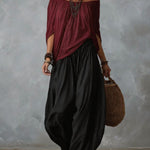 A woman models a bohemian-style outfit featuring a draped off-the-shoulder top in deep burgundy and wide-leg, voluminous black pants. She is accessorized with layered silver necklaces, bracelets, and large hoop earrings, and carries a woven brown tote bag against a textured gray studio backdrop.