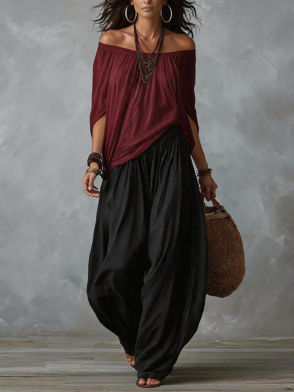 A woman models a bohemian-style outfit featuring a draped off-the-shoulder top in deep burgundy and wide-leg, voluminous black pants. She is accessorized with layered silver necklaces, bracelets, and large hoop earrings, and carries a woven brown tote bag against a textured gray studio backdrop.