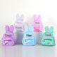 Five personalized bunny backpacks in pastel gingham checks, arranged on white display blocks against a white background. The backpacks feature rabbit ears on the back, a front zip pocket, and custom embroidered names: Daria, Elissa, and Kamila.