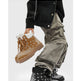 A person wearing black and gray bandana-patterned lace-up boots with gray cargo pants and a black jacket, holding a pair of tan bandana-patterned lace-up boots against a light gray background.