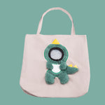 Beige tote bag with a green dinosaur plush toy on a teal background