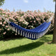 A blue hammock with white tassels is suspended between two trees in a garden setting, with green grass, a tree trunk, and a dense background of pink flowers under a blue sky.